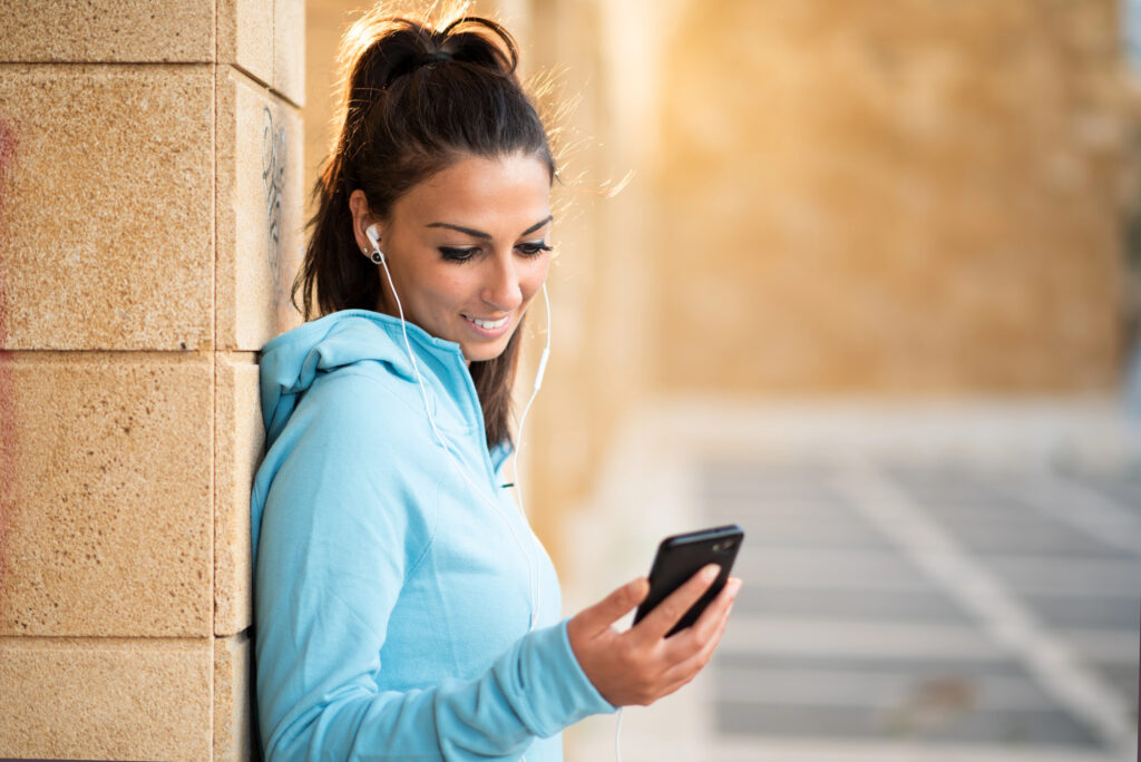 A person wearing a light blue hoodie leans against a tan brick wall, smiling at their smartphone. Earphones in, they enjoy pocket coaching with dr yam the keep it simple coach outdoors, with sunlight highlighting the relaxed, cheerful vibe.