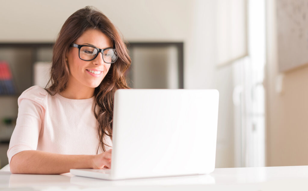 A woman with long brown hair and glasses is sitting at a white desk, smiling while using a white laptop in a bright, modern room as she listens to her favorite Pocket Coaching Private Podcast.