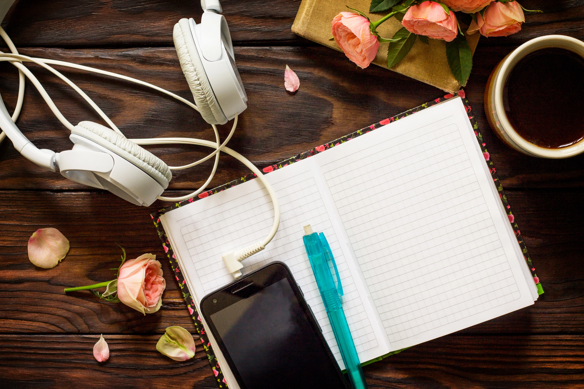 An open notebook with a blue pen, a smartphone playing a private podcast, white headphones, a cup of coffee, and pink roses rest on a dark wooden table. Some rose petals are scattered around the notebook.