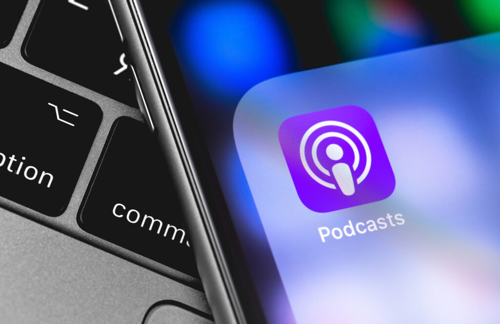 A close up of a smartphone screen displaying the Podcasts app icon next to the edge of a laptop keyboard, suggesting access to a Private Podcast or Coaching Podcast. The app icon is purple with a white symbol and the background is blurred.