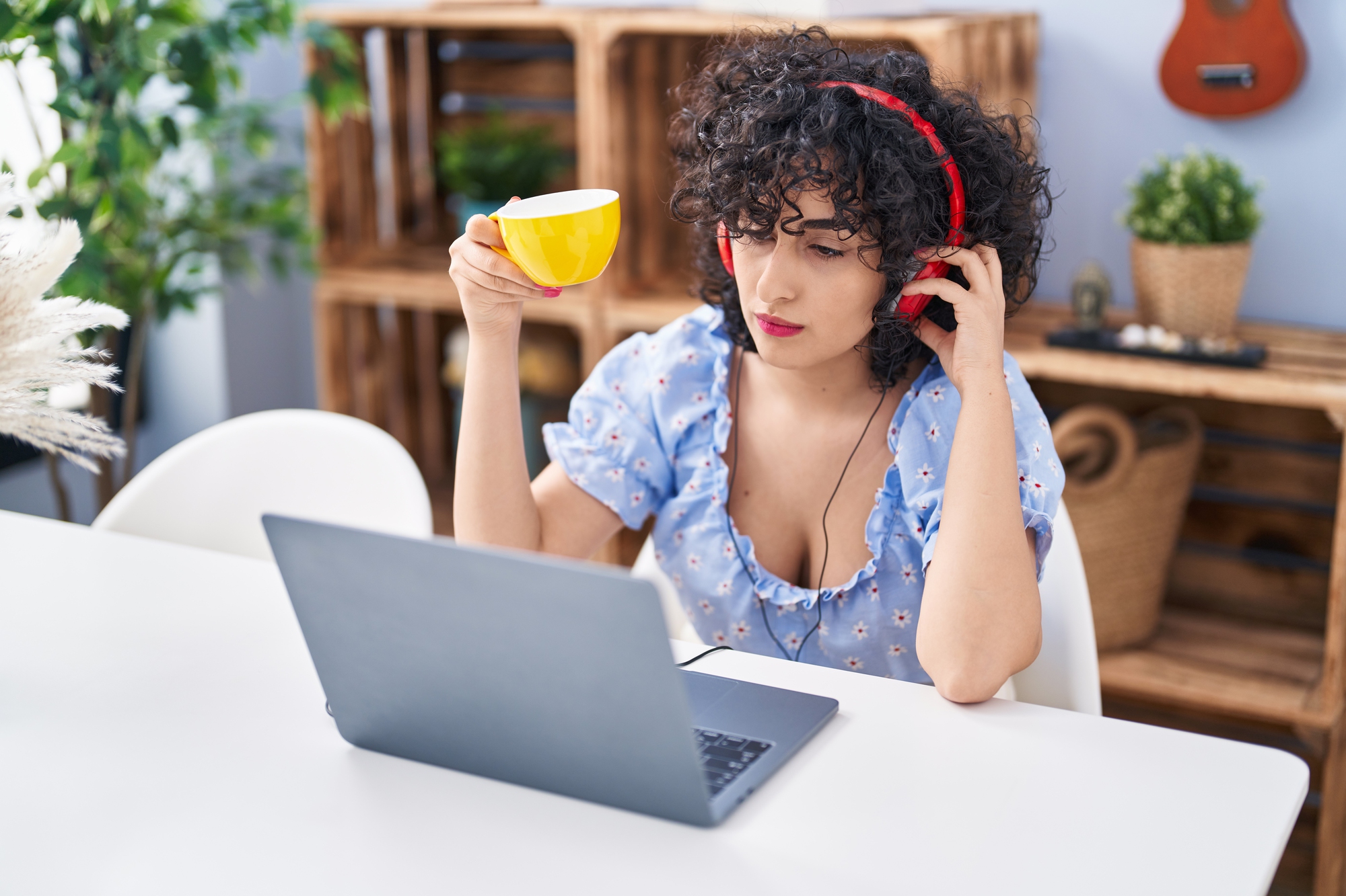 A person with curly hair wearing red headphones sits at a white table, looking at a laptop and holding a yellow mug—perhaps enjoying some pocket coaching interested in the tax season checklist. Wooden shelves and plants are visible in the background.