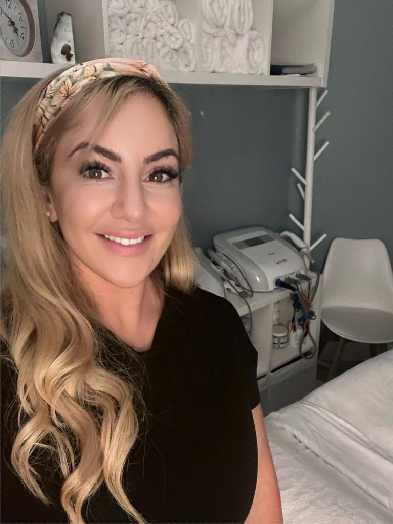 Luna - Massage Therapist is a coaching client of Keep It Simple Coach a woman with long blonde hair, wearing a black top and a headband, smiles at the camera in a room with a treatment bed, medical devices, and shelves—an inviting space for those interested in the Pocket Coaching Program.